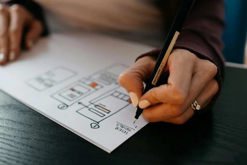 A hand drawing a wireframe on paper, one panel labelled user goals.