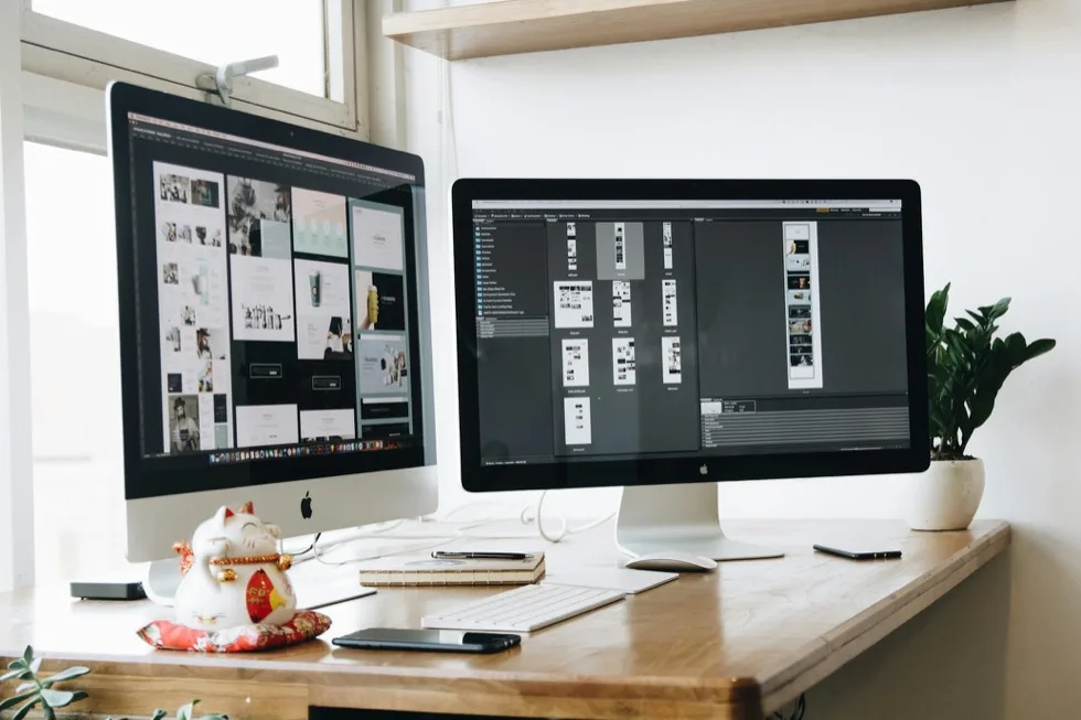 Two desktop screens on a desk, one holding a grid of page layouts and the other a folder of design files.