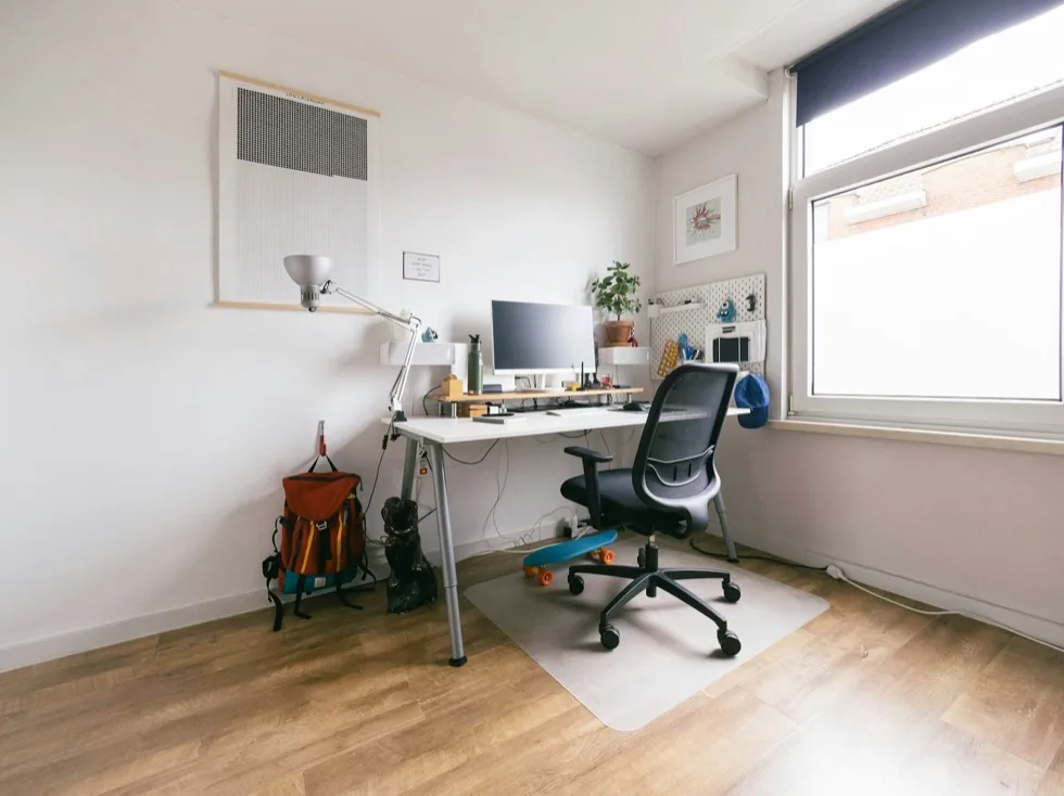 An unoccupied desk in a bright room, chair turned out, backpack and skateboard on the floor.